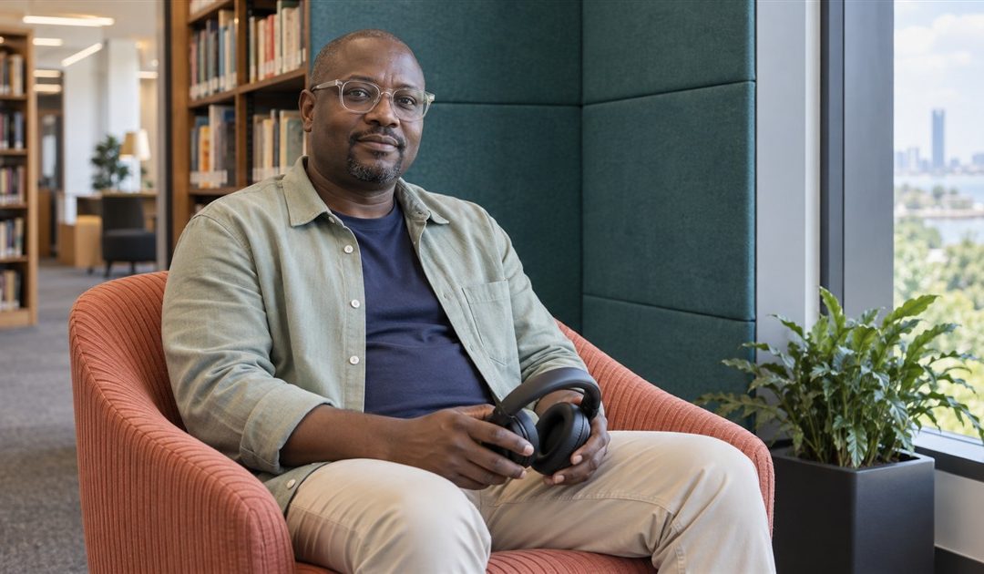 Man with glasses holding headphones in his lap while seated in a coral chair in a quiet library alcove